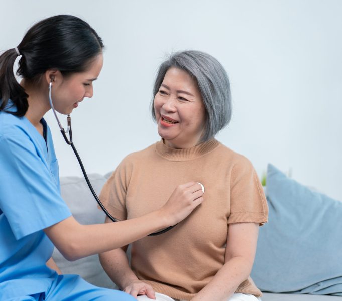 Asian nurse using stethoscope checking on heartbeat examining of senior female patient at home. Elderly care heart diseases or cardiology. Home nursing and healthcare caregiver concept